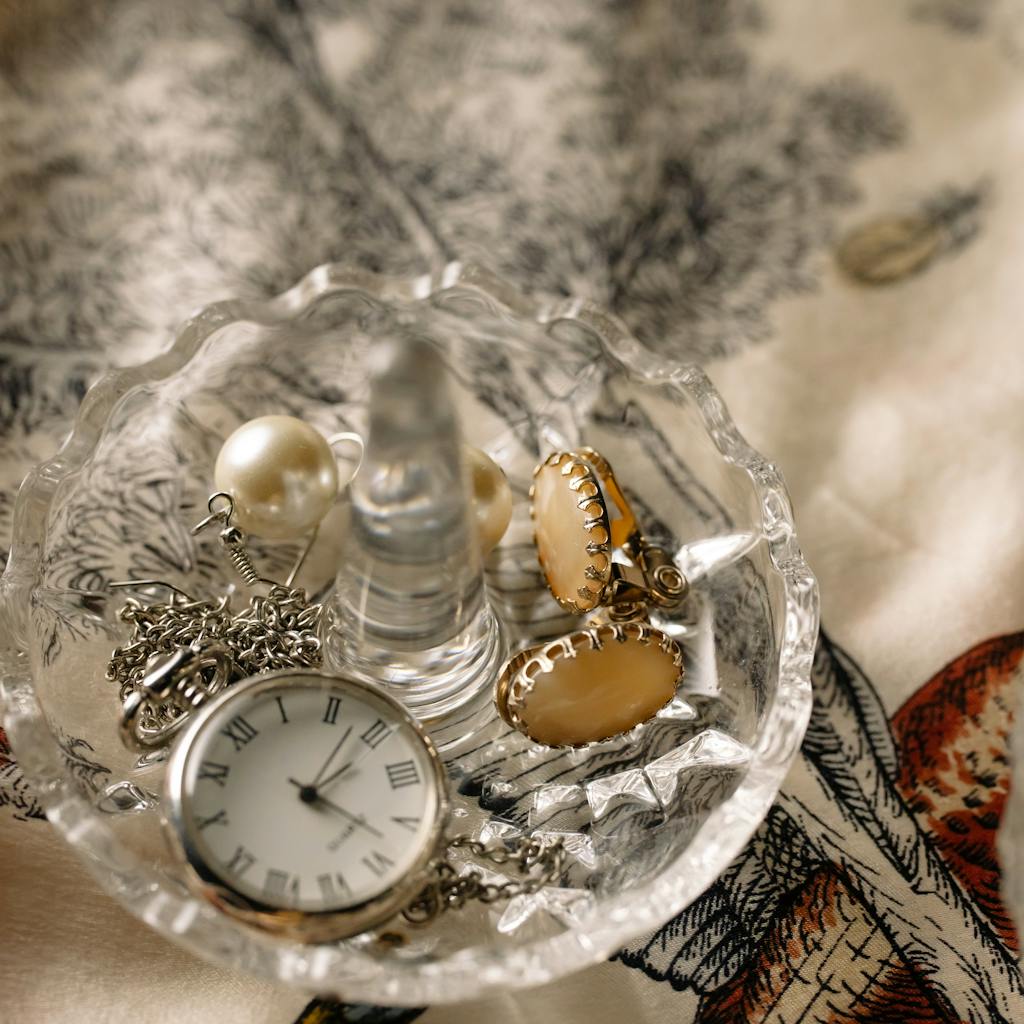 A beautiful display of jewelry and a pocket watch in a crystal dish on a patterned fabric.